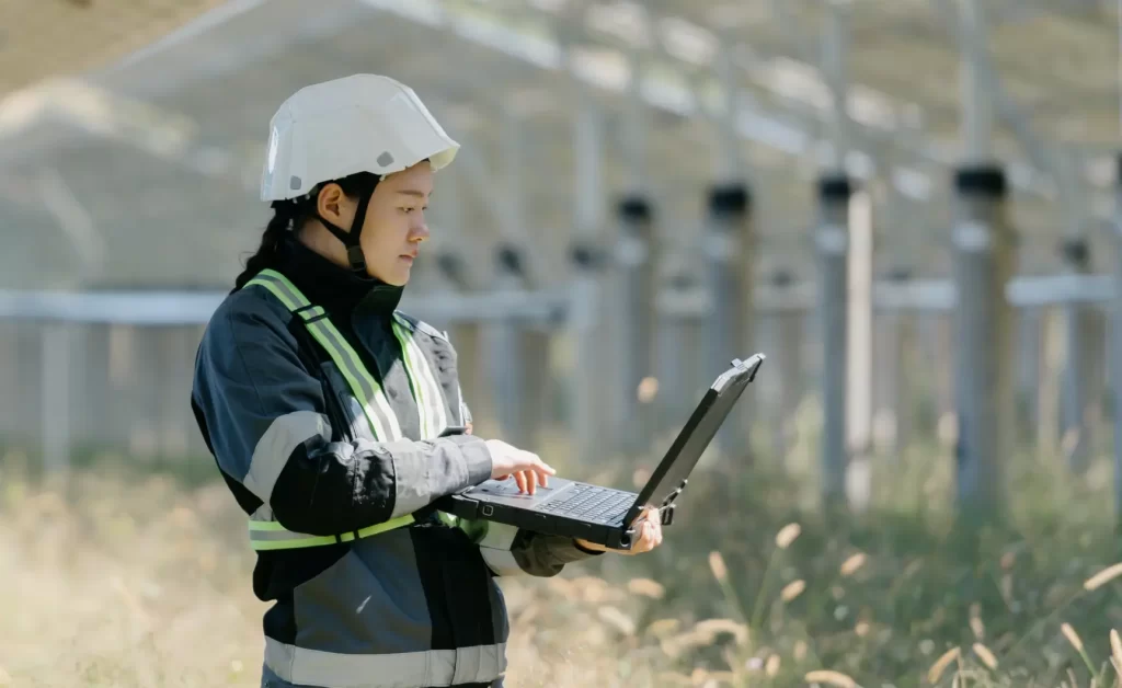 Employee next to greenhouse with laptop and hardhat to represent the blend of lean manufacturing and sustainability.