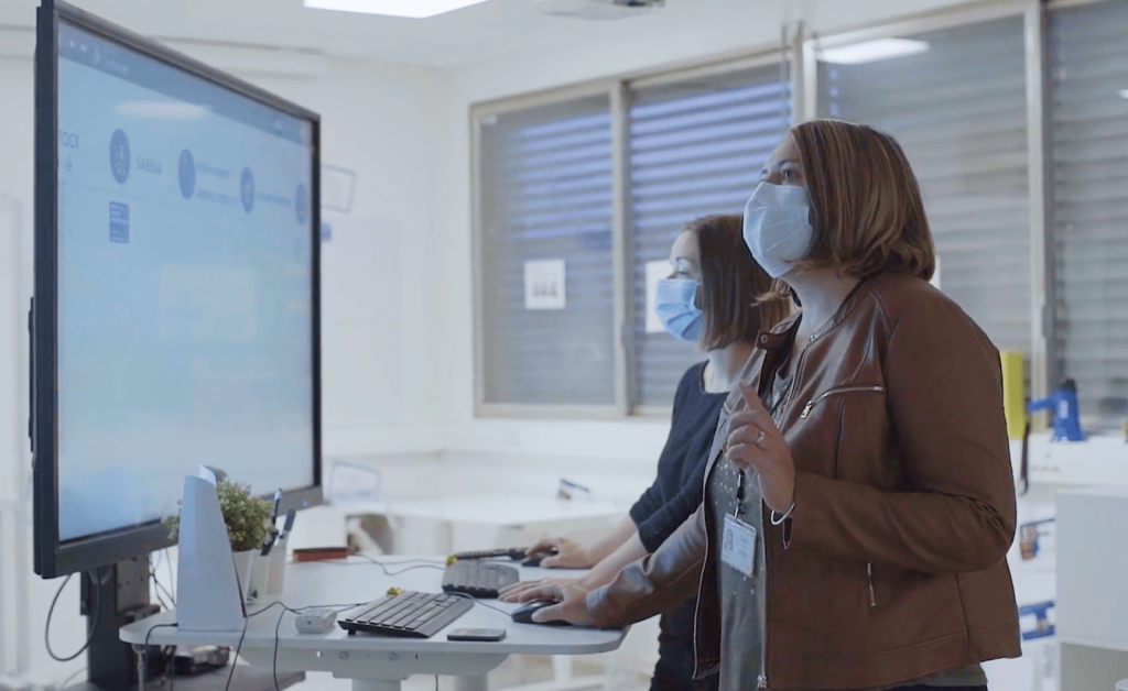 Deux femmes collaborant, debout devant un grand appareil à écran tactile utilisant iObeya dans leur environnement de bureau.