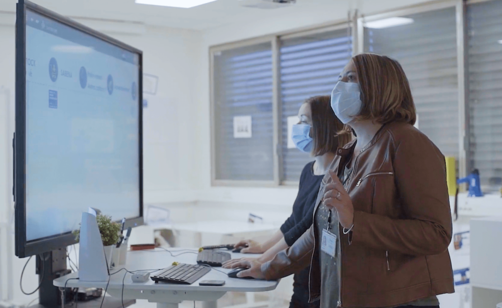 Two women collaborating, standing in front of large touchscreen device using iObeya in their office setting.