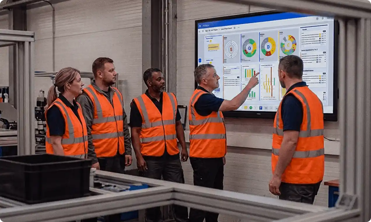 A manufacturing team in high-visibility vests gathered around a digital SQCDP board on iObeya, with a supervisor pointing to a KPI during the shift meeting